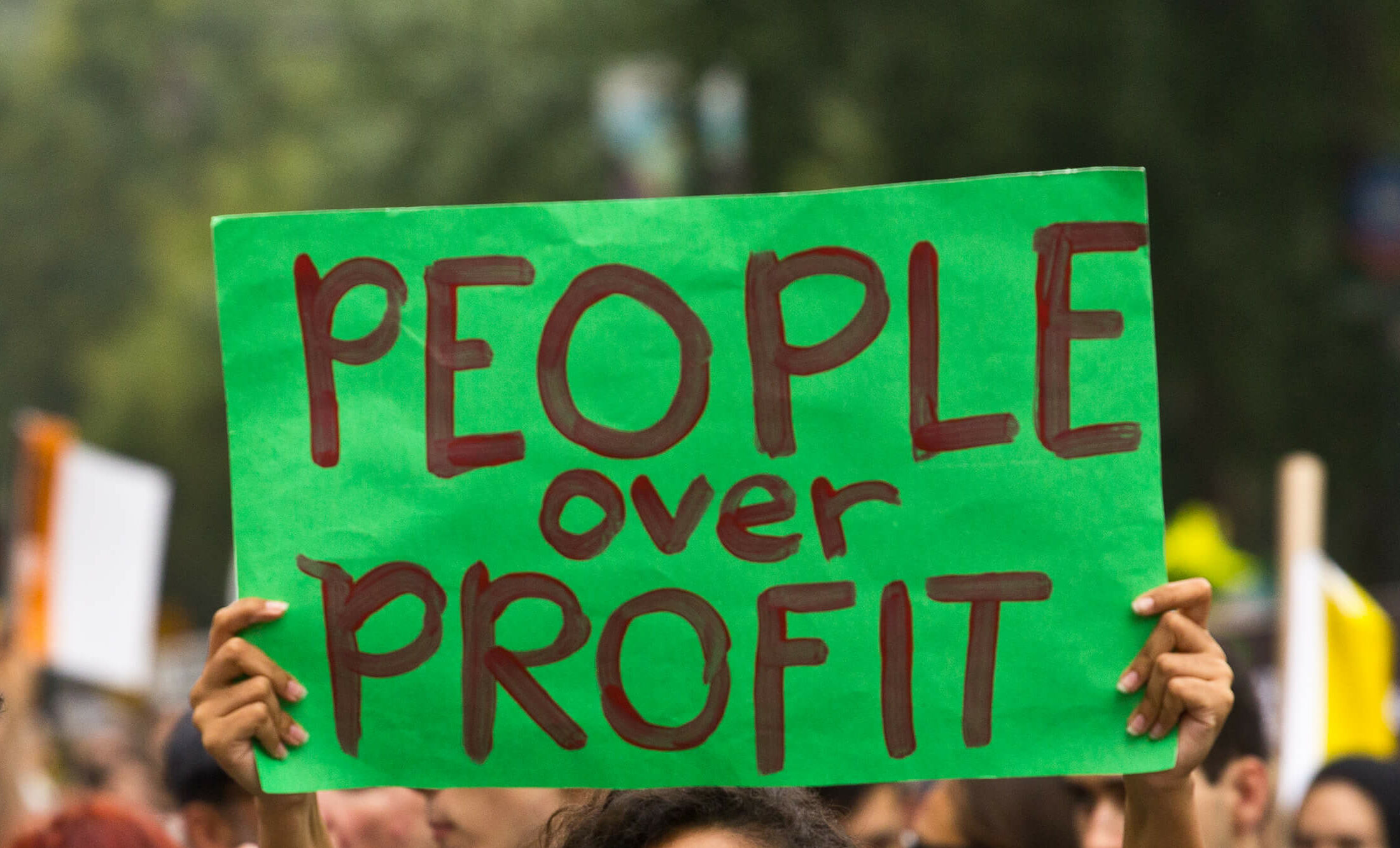 A person holding a sign during a protest. The sign reads "People over Profit"