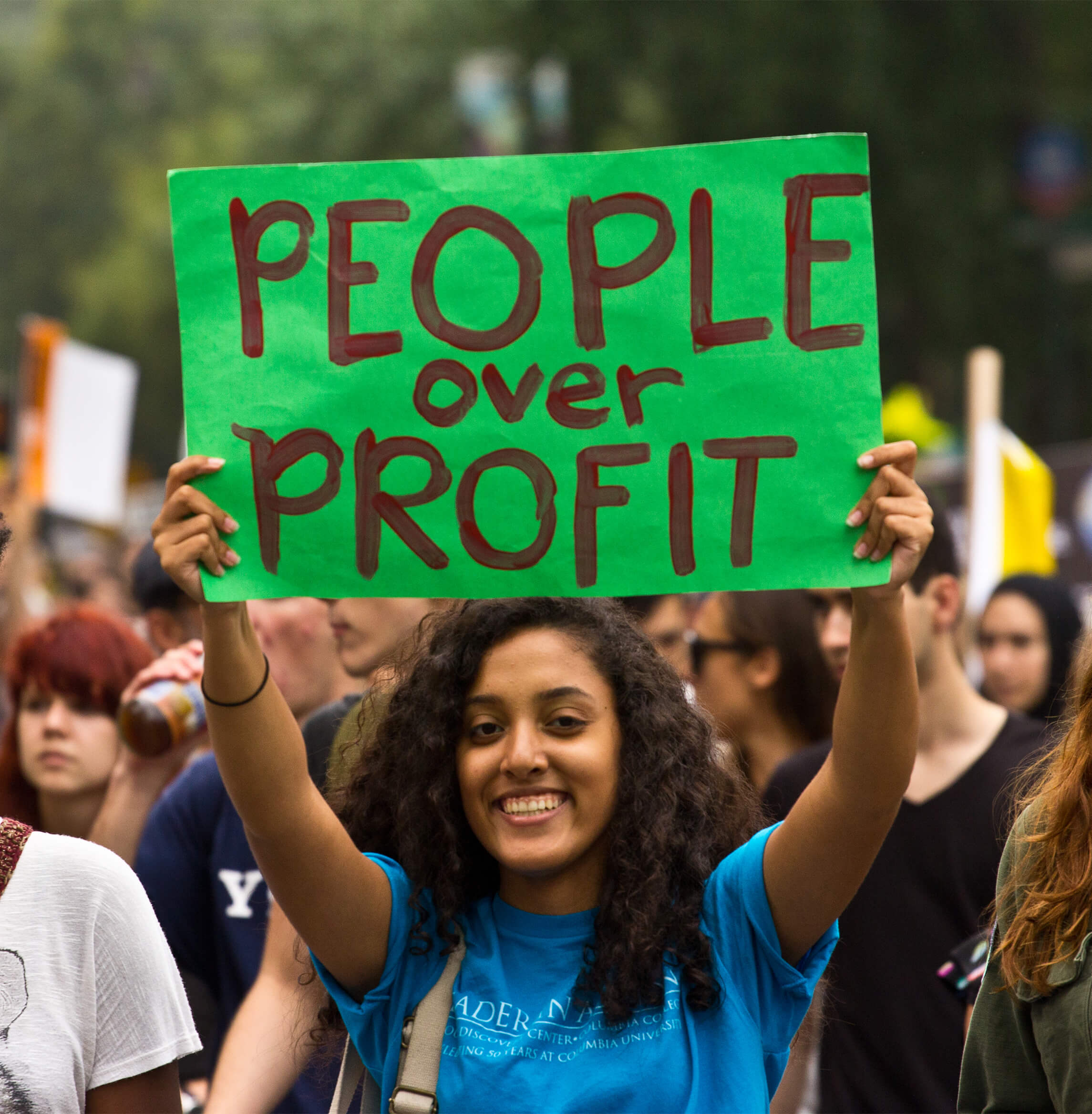 A person holding a sign during a protest. The sign reads "People over Profit"
